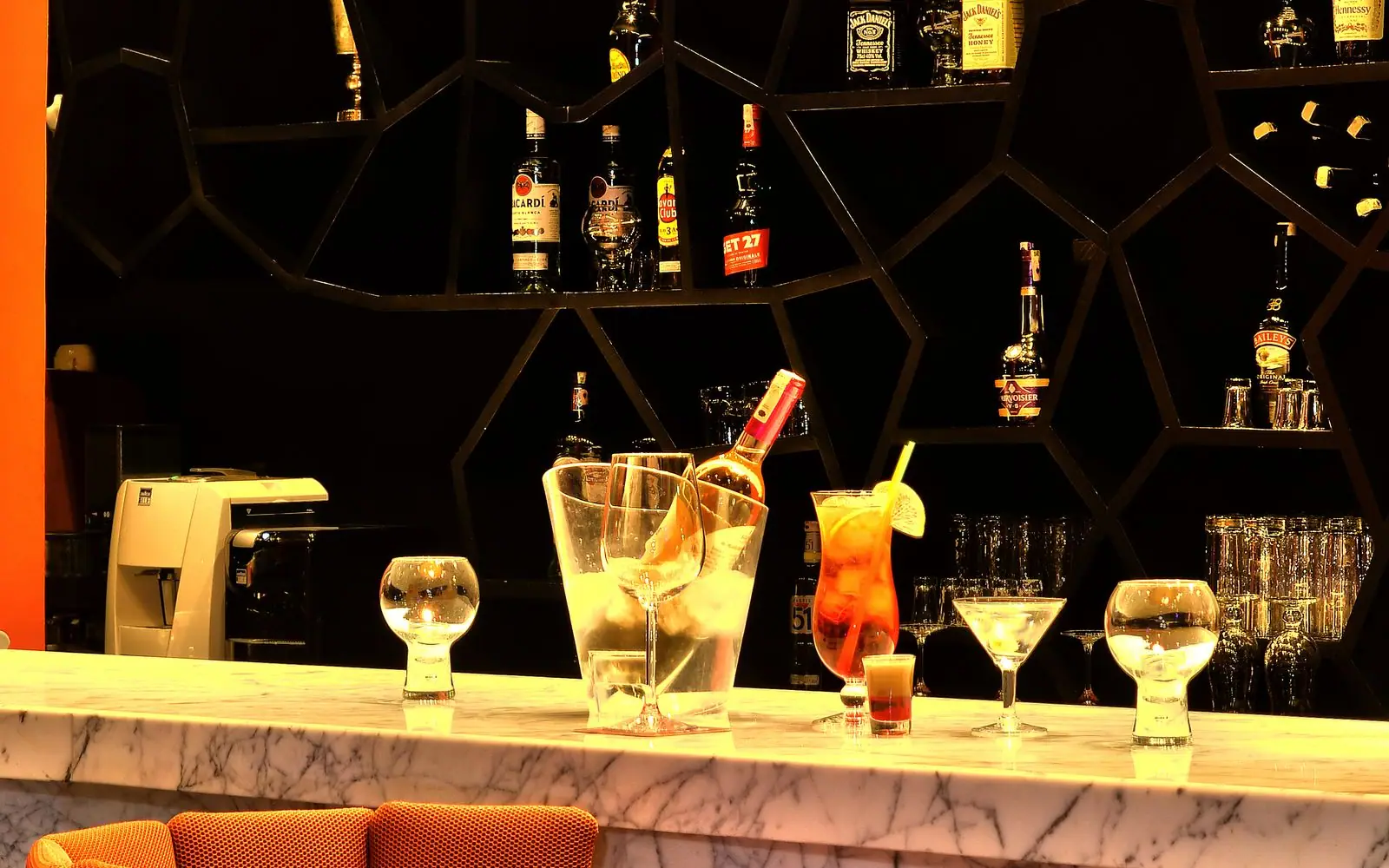 Dimly lit craft cocktail bar at night with a glowing brass back-bar of backlit liquor bottles and a row of empty velvet bar stools